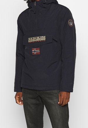 Uomo che indossa una giacca con cappuccio Napapijri blu navy con toppa della bandiera norvegese e tasca frontale, abbinata a jeans grigio scuro.