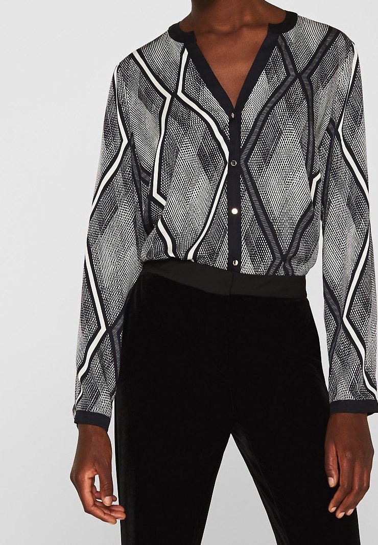 Blouse noire et blanche à manches longues à motif géométrique avec boutons, associée à un pantalon noir uni sur une personne à la peau foncée.