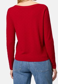 Haut rouge à manches longues avec un motif texturé de petits points, col ouvert et tissu doux. Associé à un jean en denim bleu clair.