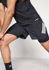 Čierne športové šortky s priedušným sieťovinovým panelom, elastickým pásom a bielymi pruhovanými akcentmi na boku. Textília pôsobí ľahkým dojmom s hladkou štruktúrou.