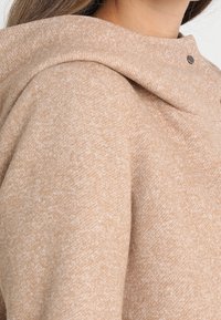 Tan hoodie med tekstur, stor omslagskrave og trykknaplukning. Blødt stof med et subtilt prikmønster.