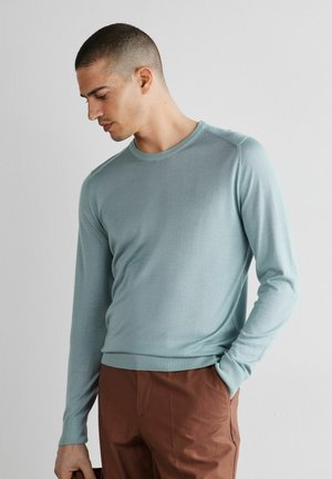 Mand med kort hår iført lyseblå langærmet sweater og brune bukser, stående med den ene hånd i lommen, kigger ned mod siden.