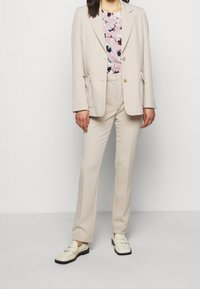 Costume beige ajusté avec un blazer à un bouton et un pantalon droit, associé à un chemisier à motifs floraux et des mocassins crème.