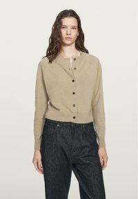 Cardigan lavorato a maglia beige con scollo rotondo, chiusura frontale con bottoni e maniche lunghe, abbinato a jeans di denim scuri. Tessuto semplice, senza motivi.