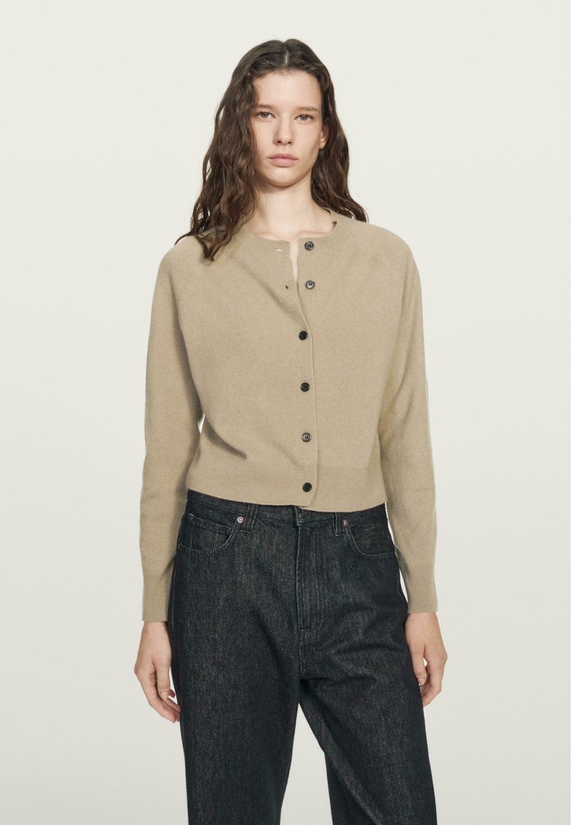 Cardigan lavorato a maglia beige con scollo rotondo, chiusura frontale con bottoni e maniche lunghe, abbinato a jeans di denim scuri. Tessuto semplice, senza motivi.