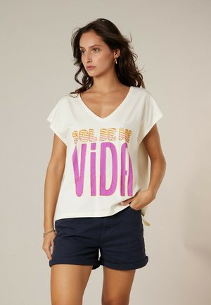 Femme aux longs cheveux foncés portant un T-shirt blanc à col en V avec le texte "sol de mi vida" en rose et orange, assorti à un short bleu foncé retroussé.