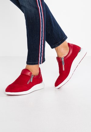 Zapatillas rojas sin cordones con cremalleras laterales, usadas con jeans azul oscuro que tienen costuras laterales a rayas en rojo, blanco y azul.