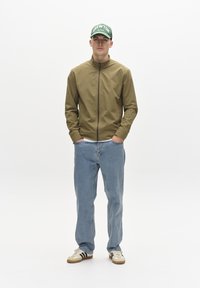 Olivgrüne Zip-Jacke, hellblaue gerade Jeans und beiges Sneaker mit schwarzen Streifen. Die Mütze hat weißen Text auf grünem Hintergrund.