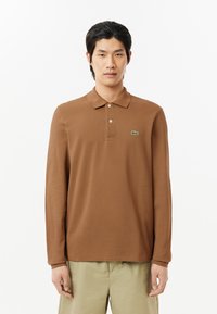 Långärmad brun polo med krage i klassisk stil, tillverkad av bomull, med en liten grön krokodil-logotyp på vänster bröst.