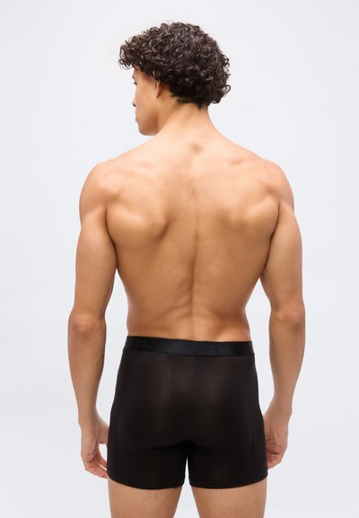 Άνδρας με σγουρά μαλλιά, εμφανιζόμενος από πίσω, φοράει μαύρα μποξέρ, σε απλό λευκό φόντο.