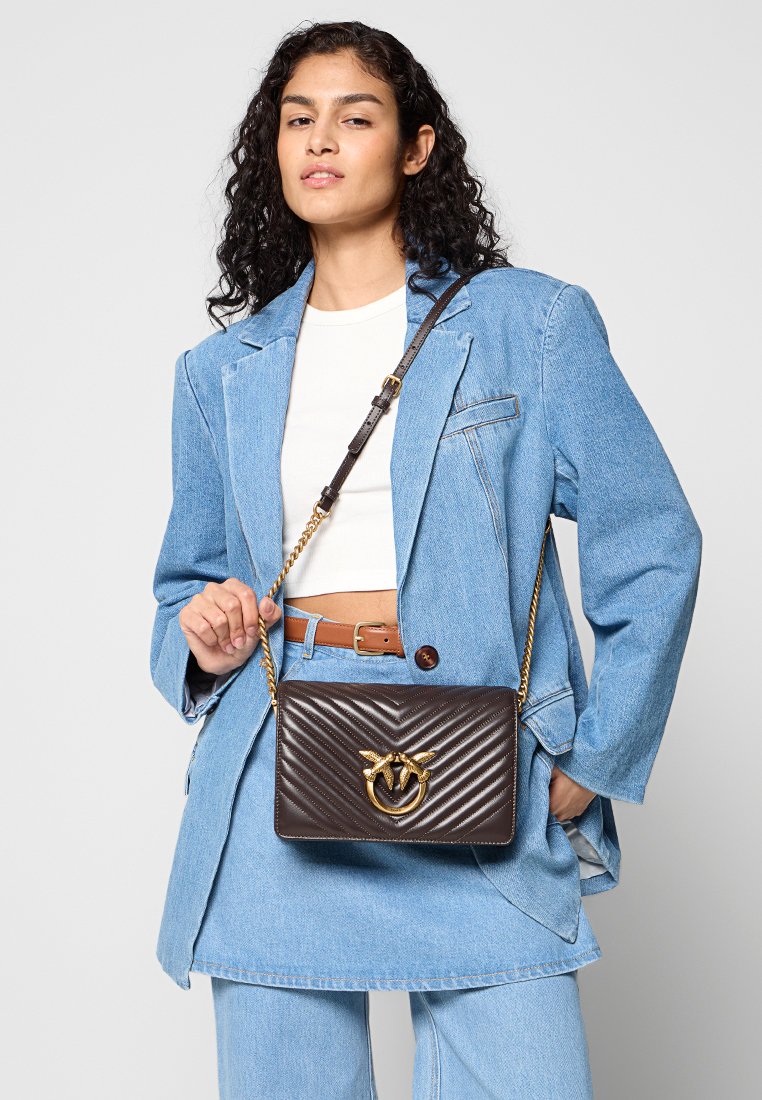 Brązowa pikowana skórzana torebka typu crossbody z złotym akcentem metalowym, wyposażona w łańcuszkowy pasek i wyróżniającym się detalem z logo.