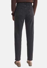 Kobieta ubrana w czarne dżinsy z wysokim stanem z tylnymi kieszeniami, brązowy sweter i taupe wsuwane buty, stojąca tyłem na białym tle.