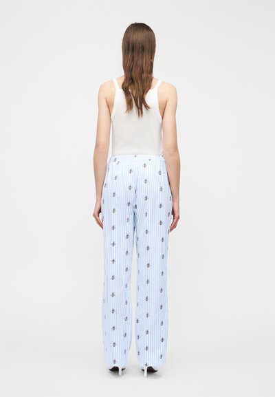 Kvinde med langt brunt hår iført en hvid tanktop og lyseblå bukser med broderede bier, stående med ryggen mod en hvid baggrund.