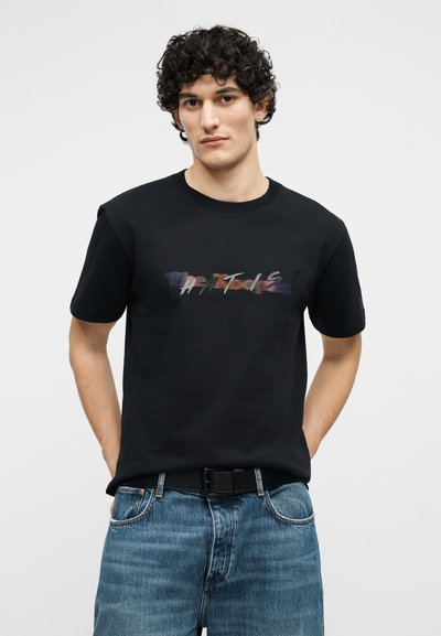 Νεαρός με σγουρά μαλλιά, που φοράει μαύρο μπλουζάκι με τύπωμα "THAT IS" και μπλε τζιν, στέκεται με τα χέρια στις τσέπες σε απλό φόντο.