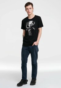 Camiseta de algodón negra con un gran gráfico de calavera blanca. El modelo lleva jeans oscuros y zapatillas negras, y está de pie contra un fondo simple.