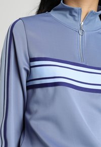 Giacca in blu chiaro con zip e colletto alto. Presenta strisce orizzontali blu e navy sulla parte anteriore. Realizzata in un tessuto liscio e elasticizzato.