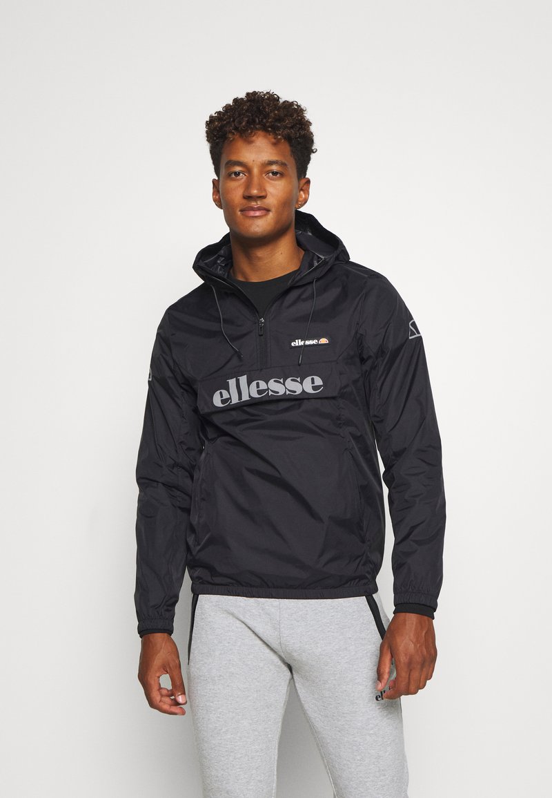 Schwarze Anorakjacke mit Kapuze, mit einem grauen Logo auf der Vorderseite und elastischen Bündchen, kombiniert mit hellgrauen Jogginghosen.