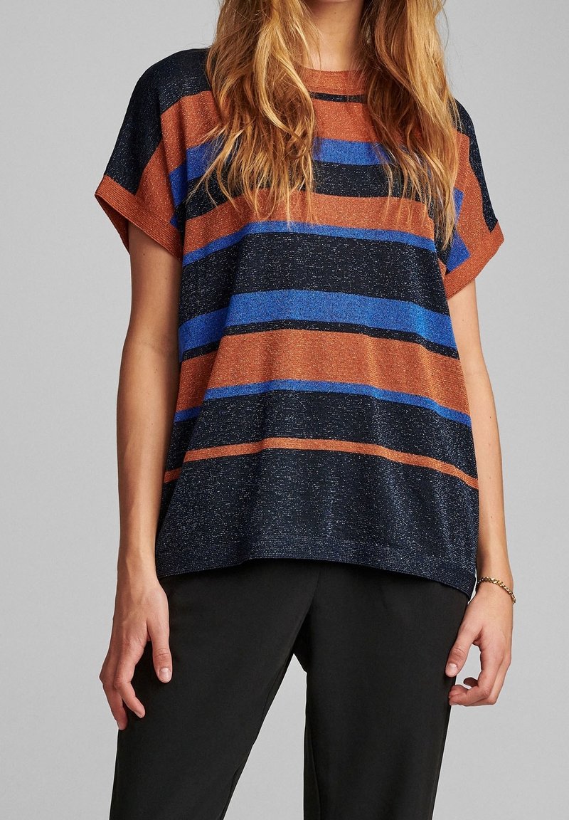 Femme portant un haut à manches courtes avec de larges rayures horizontales en rouille, bleu et noir, associée à un pantalon noir, debout devant un fond gris.