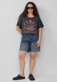 T-shirt graphique gris à manches courtes et col rond, associé à un short en denim bleu avec un ourlet effiloché et des chaussures noires à enfiler.
