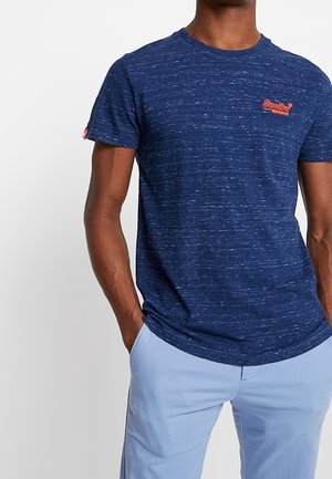 Mężczyzna w granatowym, nakrapianym T-shircie z pomarańczowym logo na piersi i jasnoniebieskich spodniach, ręce w kieszeniach, na białym tle.
