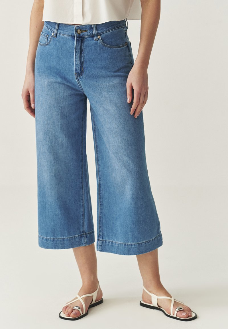 Light blue wide-leg denim jeans with a high waist, featuring a button and zip fly, front pockets, and a rolled hem. Worn with white sandals.