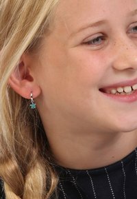 Zilveren creool met een kleine turquoise bedel in de vorm van een schildpad, gedragen door een kind met lichtblond haar en een donker gestreept shirt.