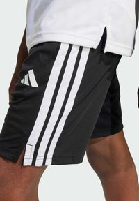 Pantalones cortos deportivos negros con rayas blancas a los lados, que presentan una textura suave, dobladillo dividido y un logo de Adidas en la parte inferior de la pierna izquierda.