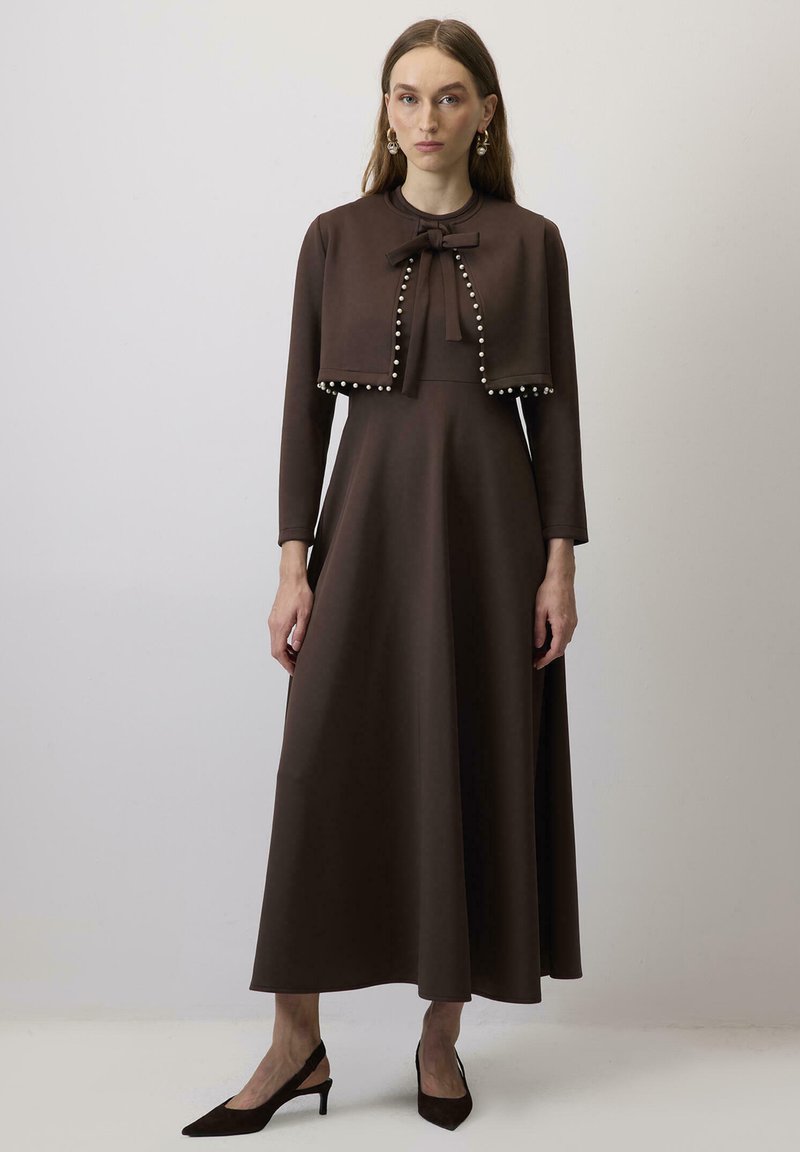 Rochie lungă maro cu mâneci lungi, bust ajustat, fustă evazată și detaliu cu fundiță. Decorațiuni cu perle albe de-a lungul tivului jachetei scurte.