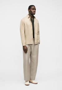 Άνδρας που στέκεται φορώντας μπεζ πουκάμισο με κουμπιά πάνω από σκούφο πράσινο πουλόβερ με V λαιμόκοψη, ανοιχτό μπεζ παντελόνι με φαρδιά μπατζάκια και λευκά αθλητικά παπούτσια με μπεζ ρίγες.