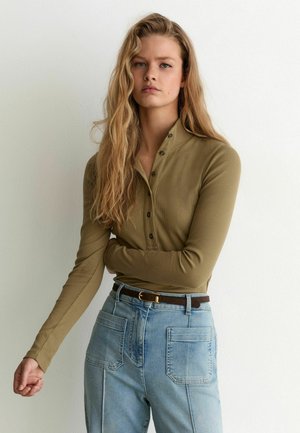 Vrouw met lang blond haar die een kaki, langärmelig, geknoopt shirt draagt en lichtblauwe, hooggetailleerde spijkerbroek met een bruine riem staat tegen een witte muur.