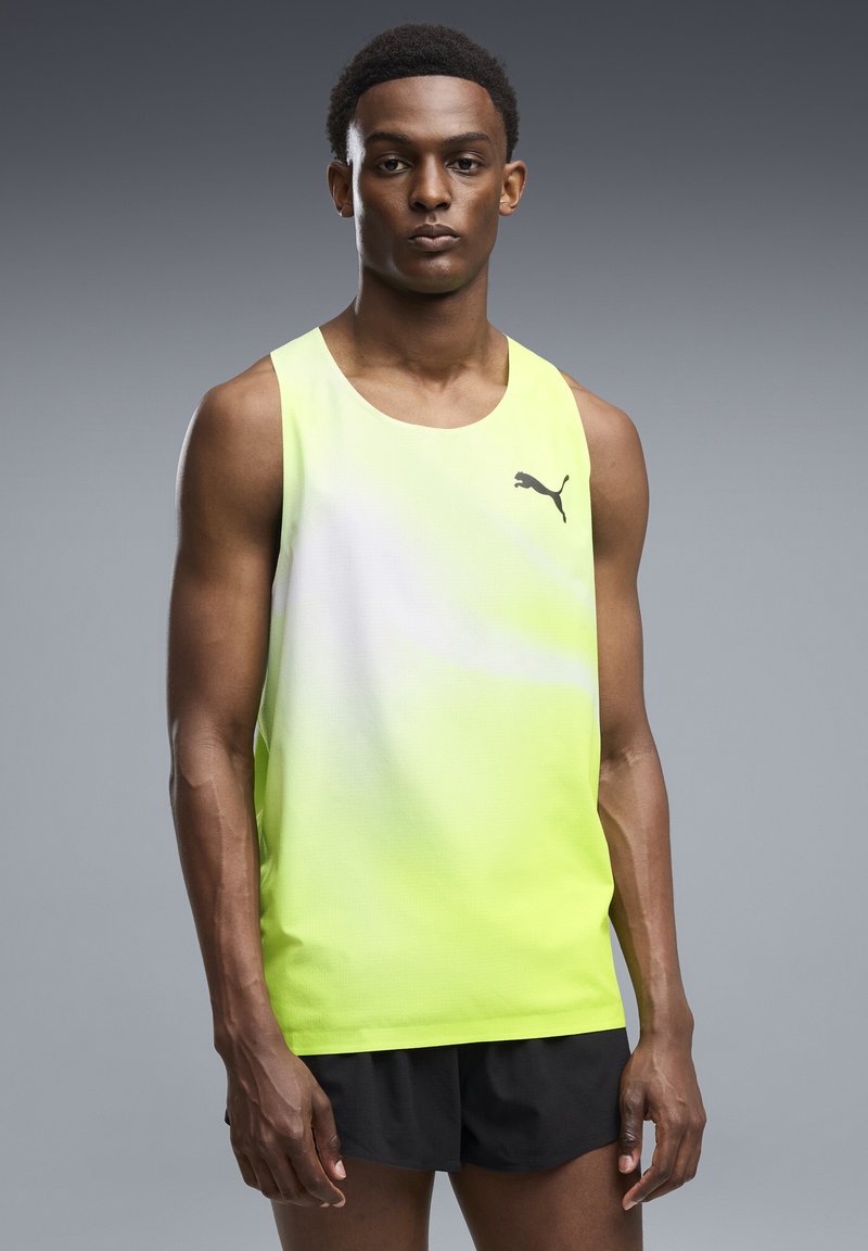 Jeune homme portant un débardeur Puma jaune et blanc et un short noir, debout devant un fond gris.