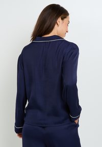 Chemise bleu marine à manches longues avec col, dotée de biais blancs le long du col et des poignets. Tissu lisse et léger avec une coupe décontractée.