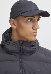 Črna izolirana jakna z visokim ovratnikom in zadrgo; teksturirana tkanina. Črna kapa s ukrivljenim šiltom. Model obrnjena rahlo levo.