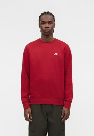 Jeune homme aux cheveux tressés portant un sweat-shirt rouge Nike et un pantalon cargo foncé, debout devant un fond clair uni.
