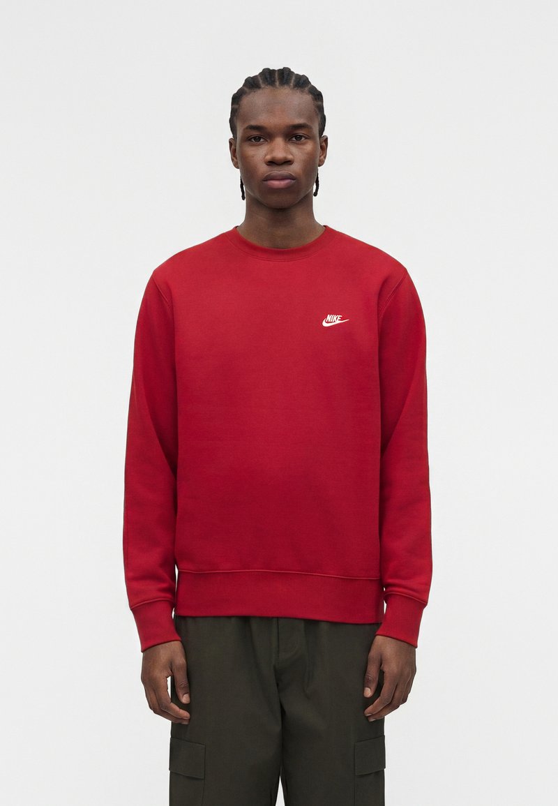 Joven con trenzas lleva una sudadera roja de Nike y pantalones cargo oscuros, de pie frente a un fondo claro y sencillo.