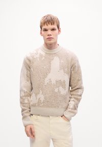 Young man with light brown hair wearing a beige and cream patterned sweater and light-colored pants, standing against a plain white background.