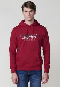 Felpa rossa in cotone con tasca a marsupio, cappuccio con coulisse e testo stampato in bianco e nero. I polsini e l'orlo a coste completano il design.