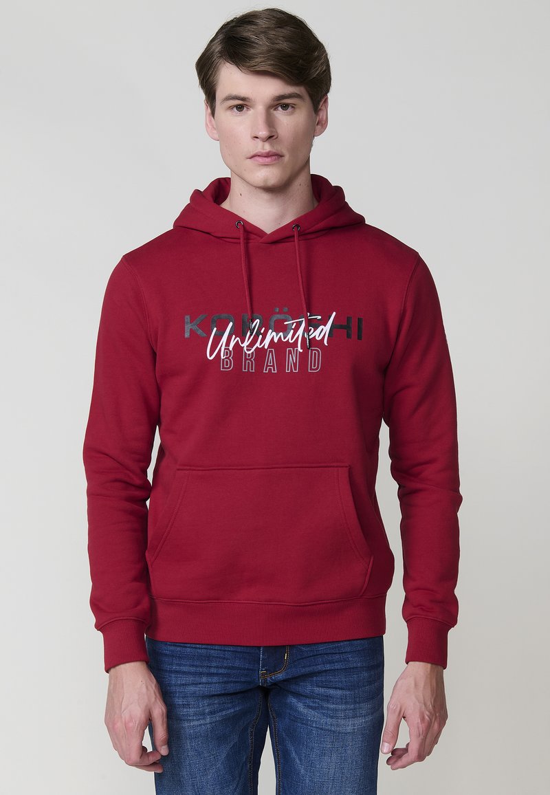 Felpa rossa in cotone con tasca a marsupio, cappuccio con coulisse e testo stampato in bianco e nero. I polsini e l'orlo a coste completano il design.