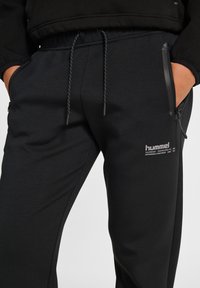 Pantalons de survêtement noirs Hummel avec une taille élastique, fermeture à cordon de serrage et poches latérales zippées. Présente un logo sur la cuisse gauche.