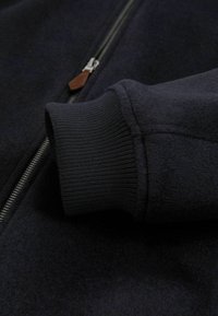 Marine blauwe zip-up jack met een gestructureerde stof, ribgebreide manchetten en een leren ritssluiting, die de stevige constructie en het moderne ontwerp benadrukt.