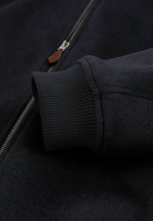 Marine blauwe zip-up jack met een gestructureerde stof, ribgebreide manchetten en een leren ritssluiting, die de stevige constructie en het moderne ontwerp benadrukt.