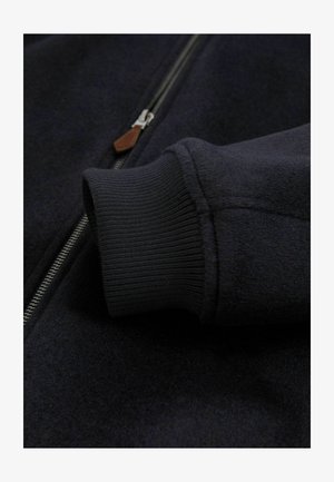 Marineblaue Jacke mit Reißverschluss aus strukturiertem Stoff, gerippten Bündchen und einem Leder-Reißverschlussanhänger, der die robuste Verarbeitung und das moderne Design hervorhebt.
