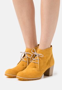 Stivali alla caviglia in suede giallo con punta arrotondata, design con lacci e tacco marrone texturizzato. Gli stivali hanno una finitura liscia e morbida.