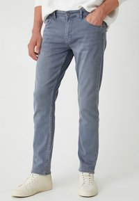 Jeans de denim de color azul claro con un ajuste slim, que presentan un diseño clásico de cinco bolsillos y cierre de botón. Llevados con zapatillas blancas.