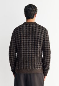 Cardigan en tricot marron à motifs avec un col montant, des poignets et un ourlet côtelés, présentant un motif à carreaux dans des nuances foncées et claires.