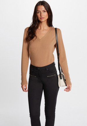 Pull en tricot beige avec un col en V et des poignets côtelés, doté de boutons sur les manches. Assorti à un jean skinny noir et un petit sac bandoulière.