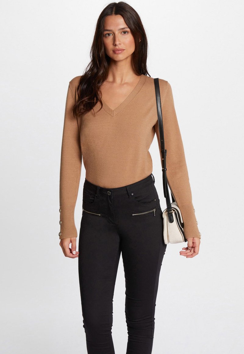 Beige gebreide trui met een V-hals en geribbelde manchetten, met knopen op de mouwen. Gecombineerd met zwarte skinny jeans en een kleine schoudertas.