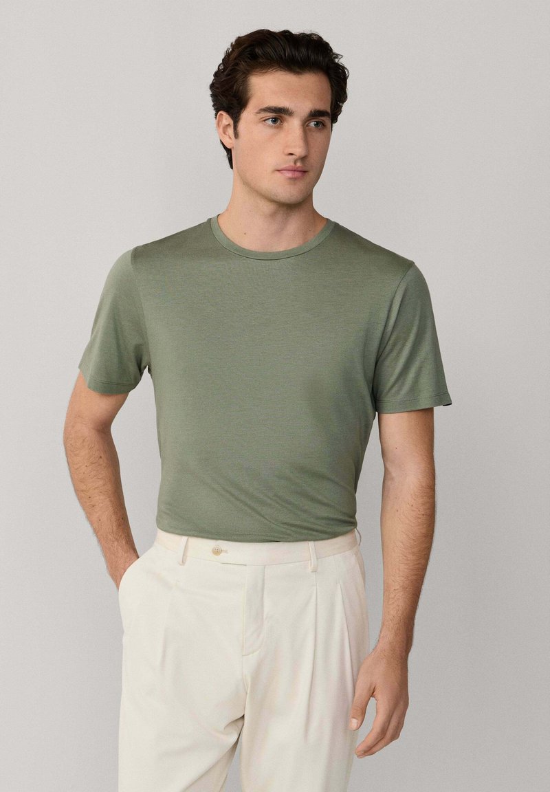 Mann trägt ein eng anliegendes olivgrünes kurzärmeliges T-Shirt, das in hoch taillierte cremefarbene plissierte Hosen gesteckt ist, posiert mit einer Hand in der Tasche.