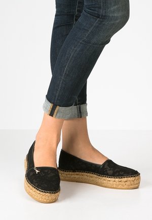 Pieds portant des espadrilles en dentelle noire avec des semelles épaisses en jute tressée, associées à un jean skinny bleu foncé avec revers, sur fond blanc.