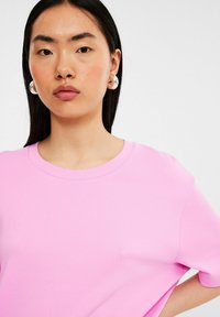 Lyserød bomulds T-shirt med rund hals, korte ærmer og en glat tekstur. Modellen bærer store, runde øreringe.
